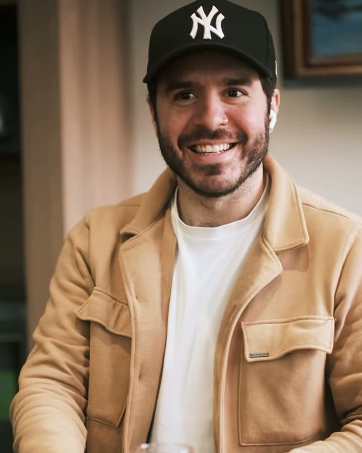 Bruno Caldas sorrindo no balcão da CINC, de boné e jaqueta caramelo